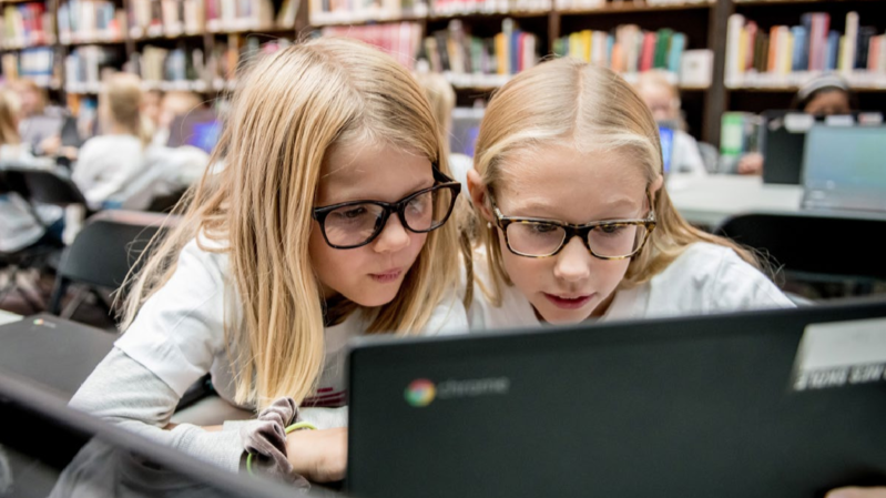 forside av bibliotekstrategien. To jenter ser på en PC skjerm i biblioteket